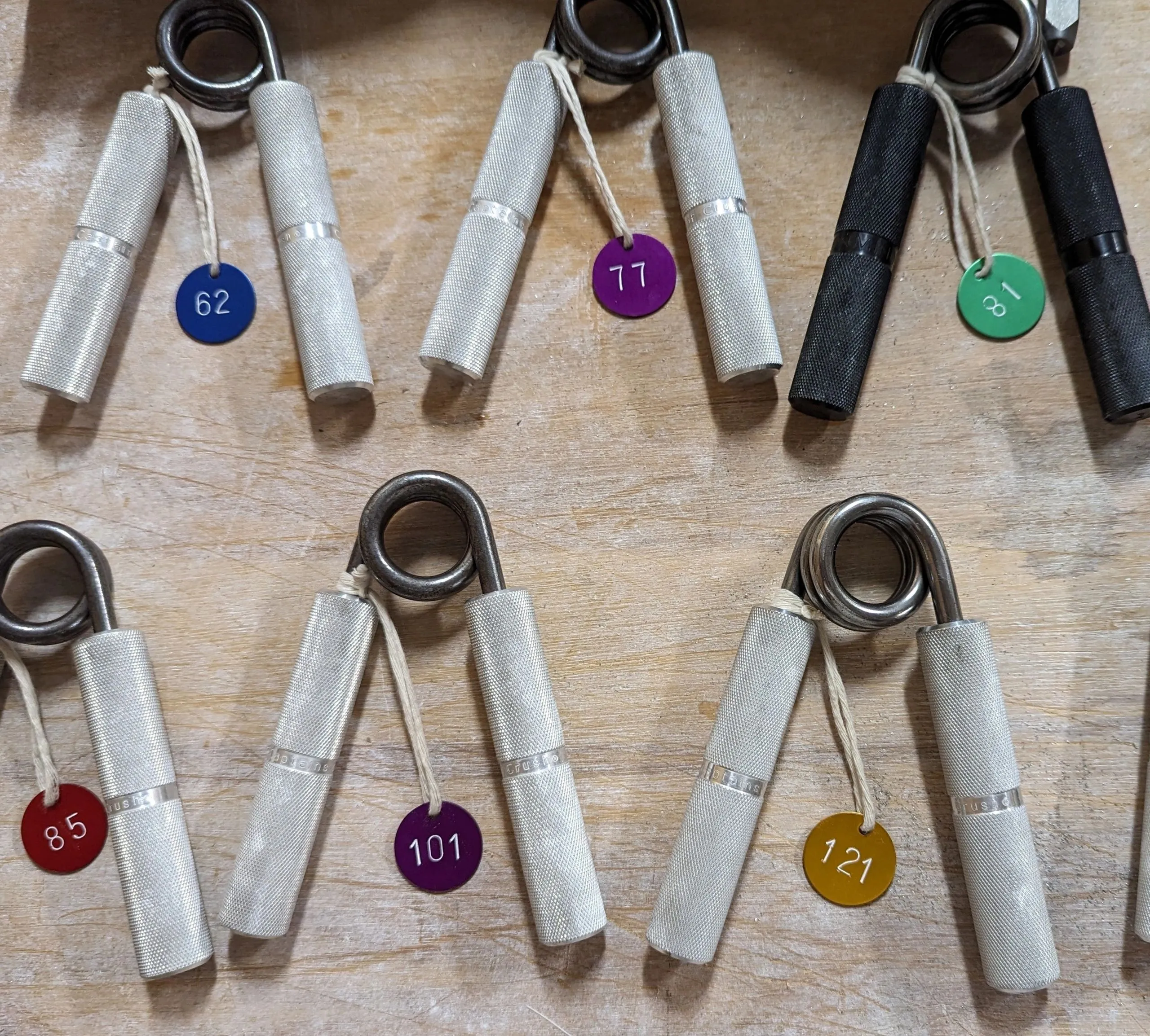 Hand grippers arrayed with color-coded RGC rating tags: 62, 77, 81, 85, 101, 121 lbs